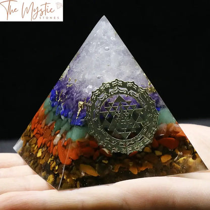 A clear pyramid embedded with layers of colorful stones representing the 7 chakras, set on a smooth base.