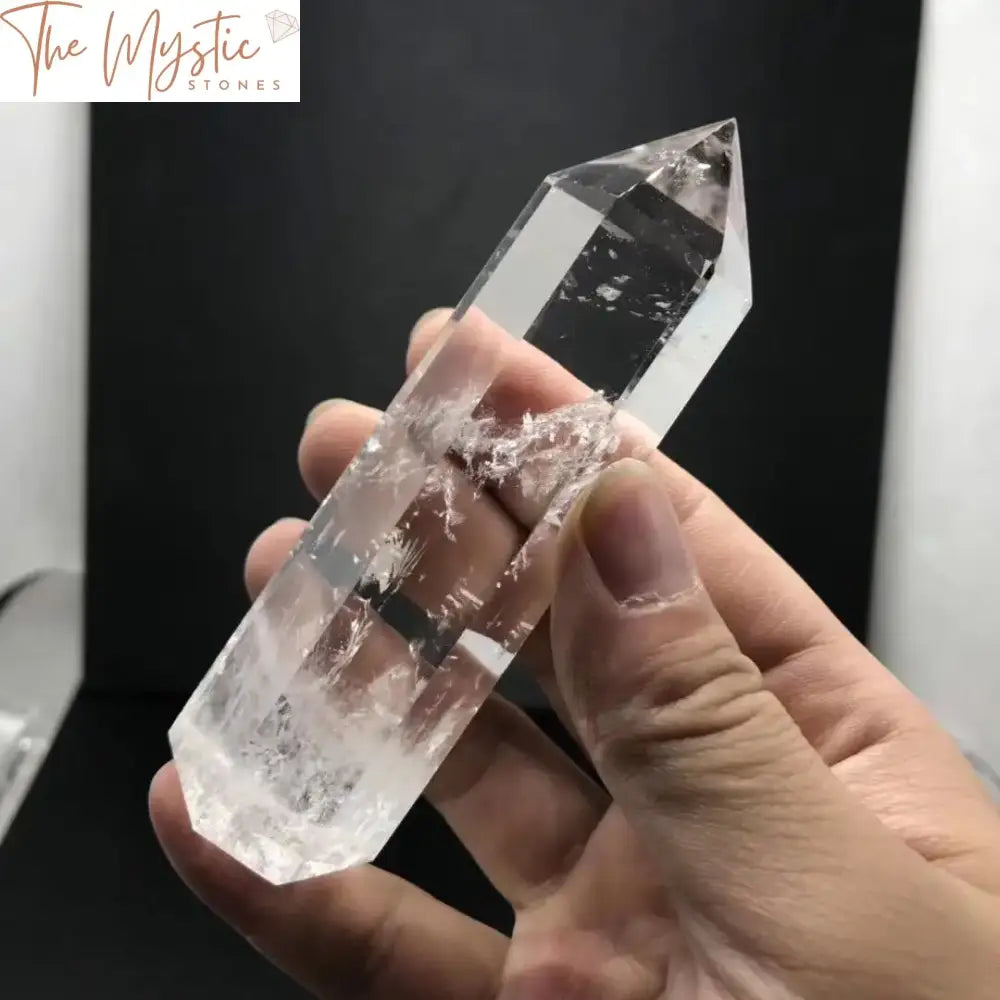 A clear, hexagonal crystal quartz obelisk with pointed ends stands upright, showcasing its transparent and glossy surface.